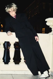 A young man with blonde hair and face tattoos wearing a black coat leaning against a stone balustrade