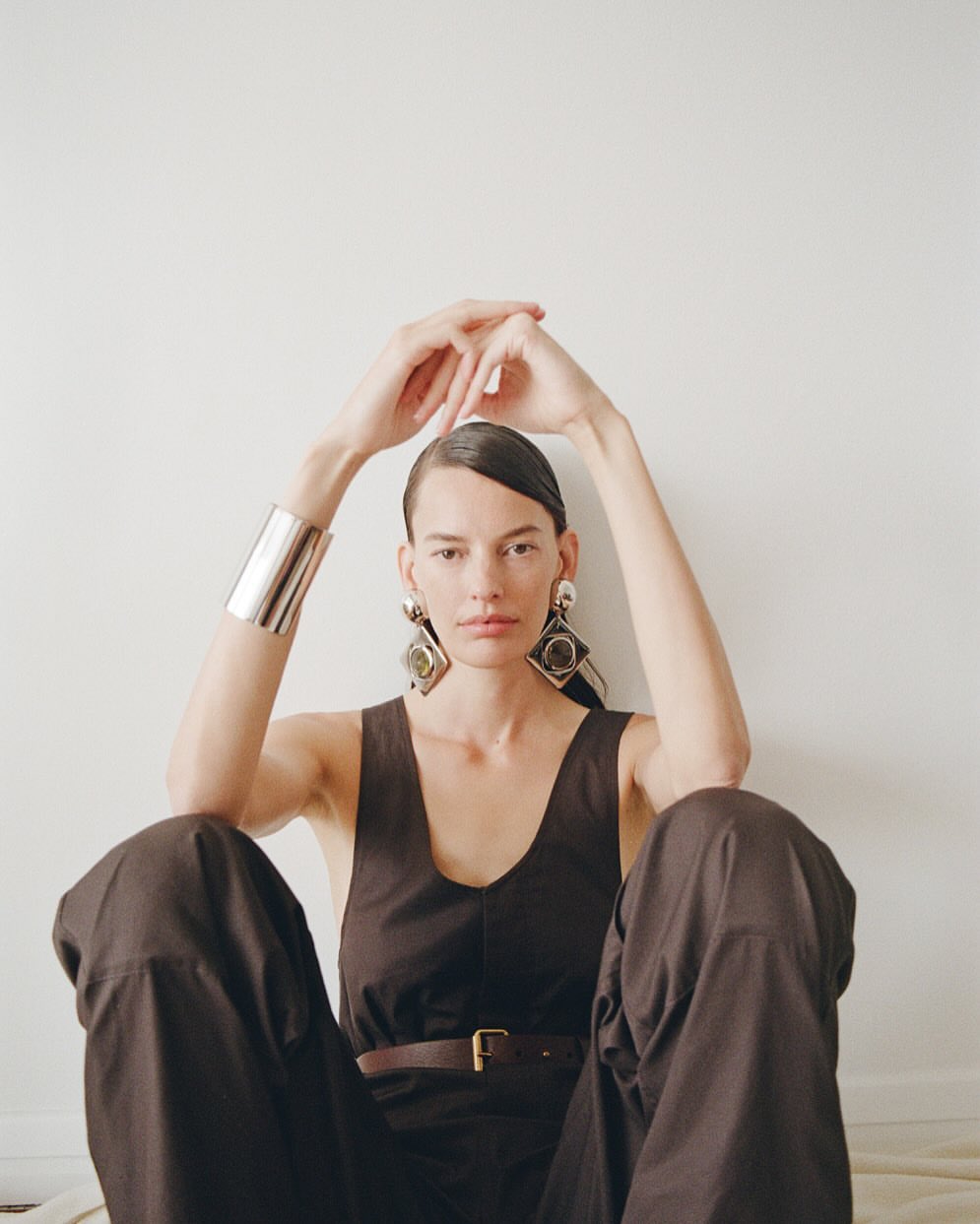 Model wearing dark sleeveless top and oversized earrings with hands clasped above head against a plain white background