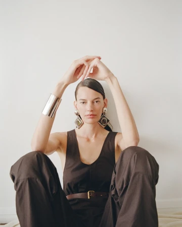 Model wearing dark sleeveless top and oversized earrings with hands clasped above head against a plain white background