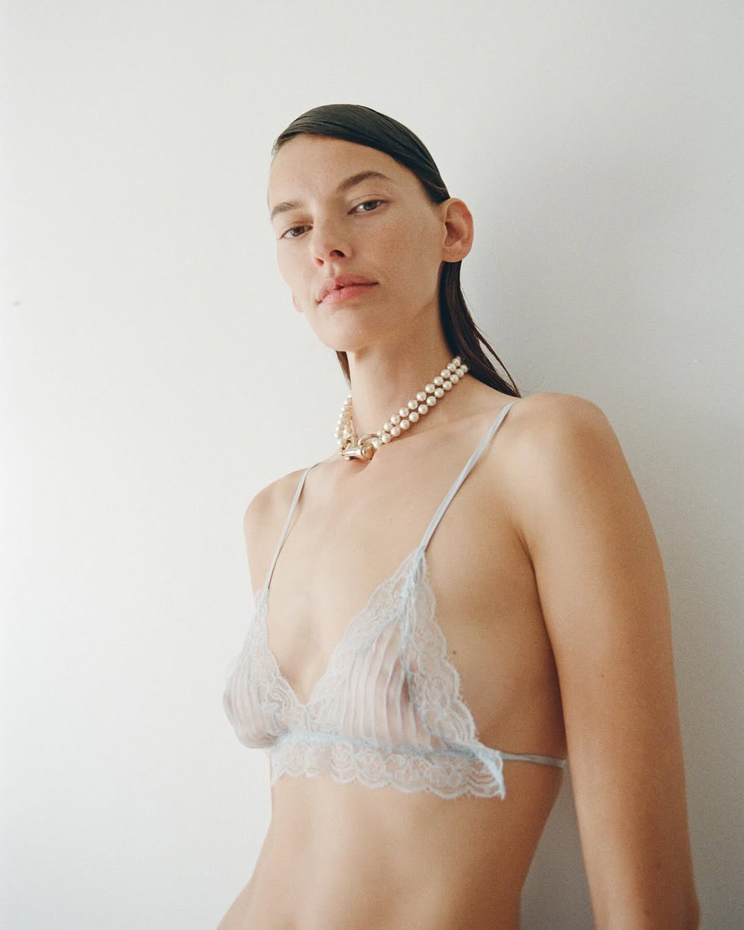 Model wearing a light blue lace bralette and a double-strand pearl necklace against a plain white background