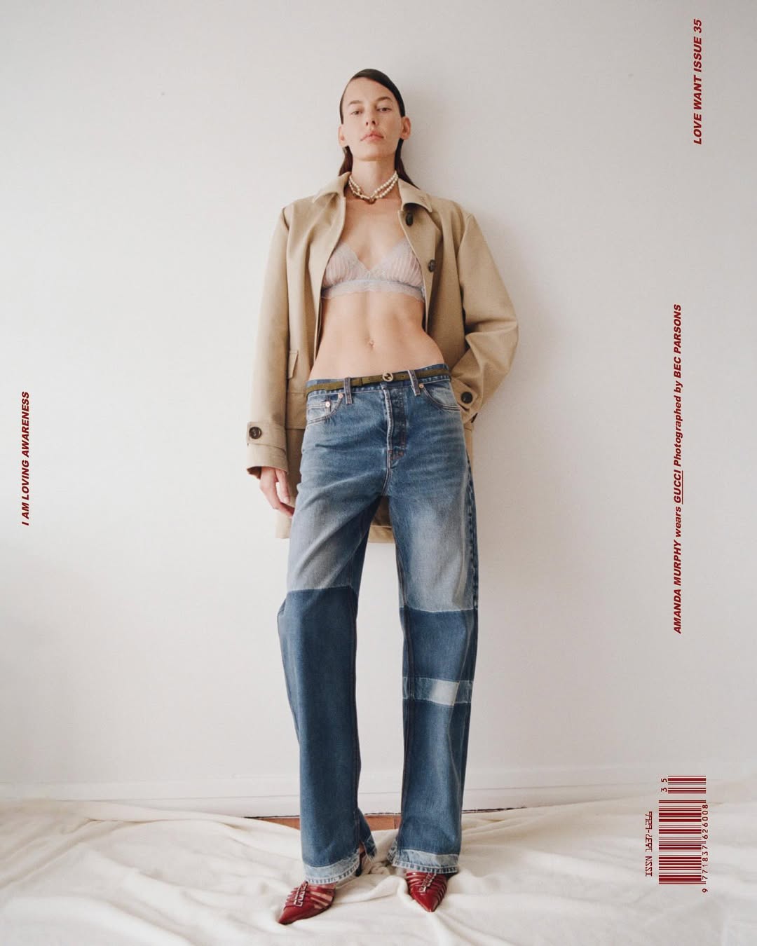 Full-length portrait of a model wearing a tan trench coat, bralette, and patchwork denim jeans against a plain white studio wall.