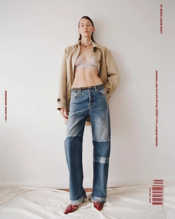 Full-length portrait of a model wearing a tan trench coat, bralette, and patchwork denim jeans against a plain white studio wall.