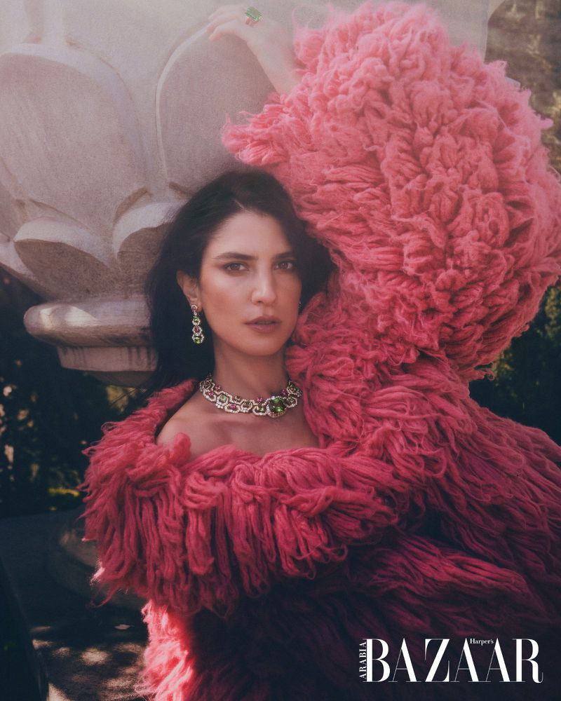 A woman with dark hair poses in a pink, textured shawl and ornate jewelry in a close-up shot outside near a stone structure.