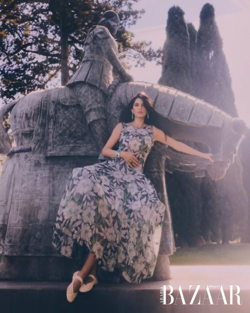 Elegant adult female in a floral gown poses next to a stone equestrian statue in a sunlit outdoor setting, fashion editorial for Bazaar.