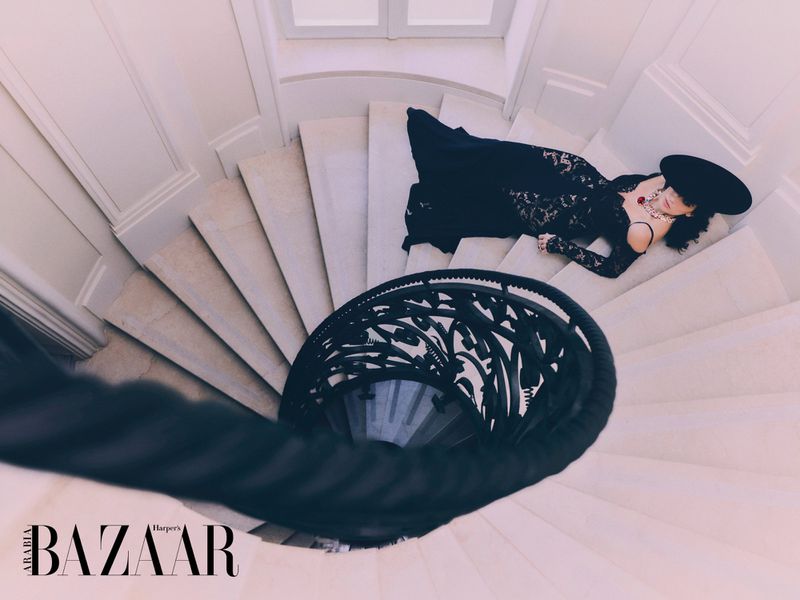 High-angle shot of a woman in a black lace dress and large hat lying on a spiral staircase in a white architectural setting.