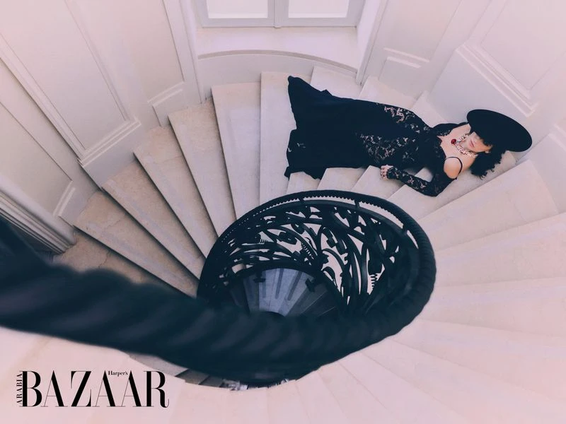 High-angle shot of a woman in a black lace dress and large hat lying on a spiral staircase in a white architectural setting.