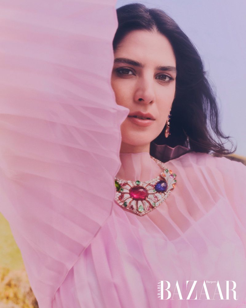 A woman in a flowing pink dress and statement necklace poses against a light sky for a Harper's Bazaar fashion editorial.