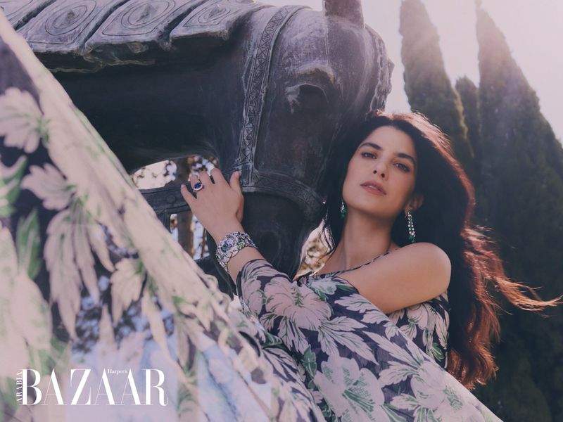 Elegant woman with long, dark hair and floral dress posing with a stone horse sculpture outdoors, accessorized with jewelry; editorial fashion