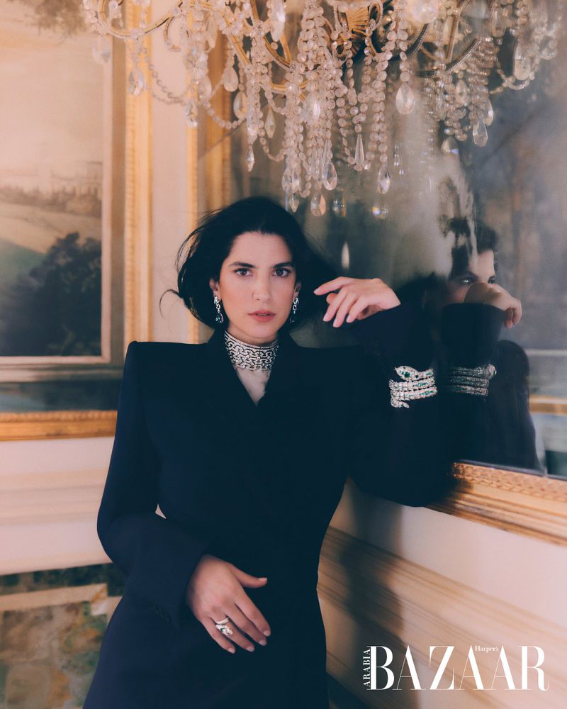 Elegant adult woman poses indoors wearing a dark dress and diamond jewelry, beneath a chandelier and ornate mirror, Harper's Bazaar logo visible.