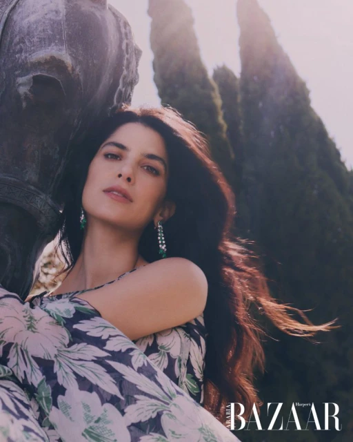 Portrait of brunette woman in floral-print dress with sculpture and trees in the background. She's wearing drop earrings and looking at camera.
