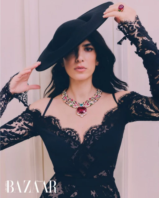 Elegant woman in black lace dress and large hat poses with statement necklace and ring indoors, as featured in Harper's Bazaar.