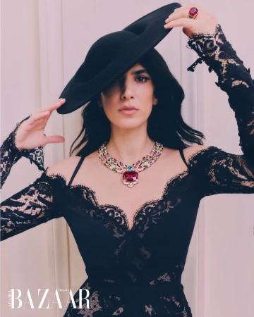 Elegant woman in black lace dress and large hat poses with statement necklace and ring indoors, as featured in Harper's Bazaar.