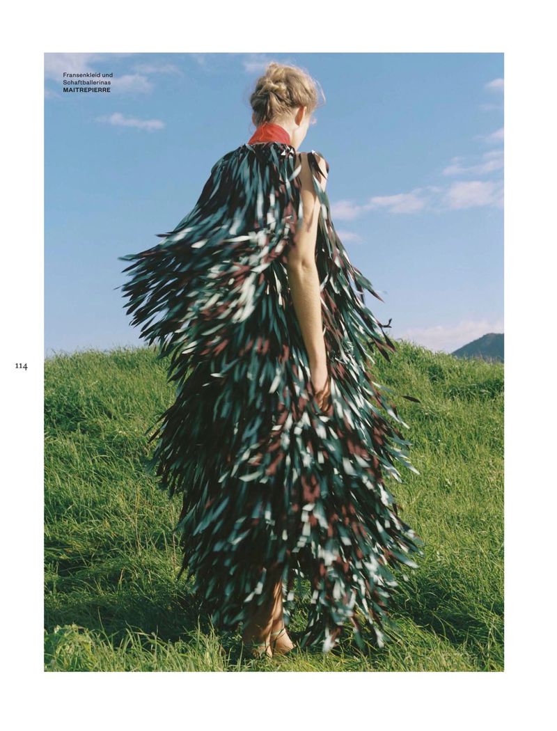 Model standing on a grassy hill wearing a textured fringed garment against a blue sky with scattered clouds
