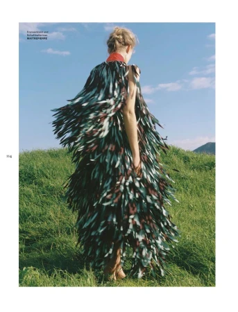 Model standing on a grassy hill wearing a textured fringed garment against a blue sky with scattered clouds