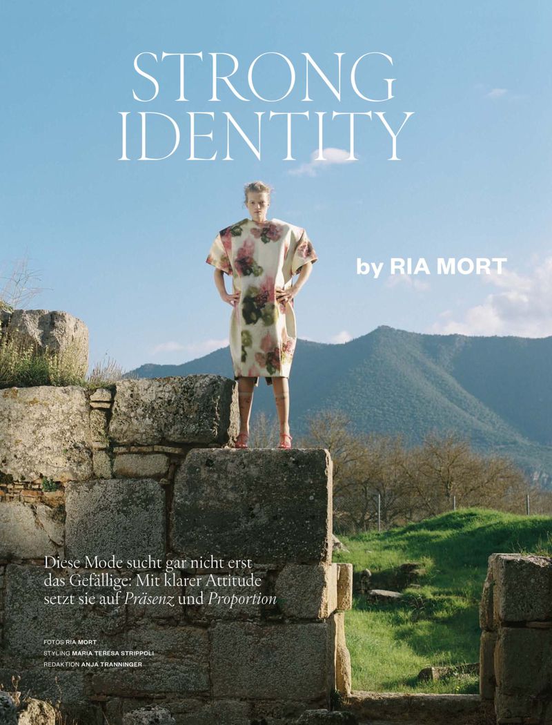 Fashion editorial image featuring a model in a floral dress standing on ancient stone ruins against a backdrop of mountains and blue sky.