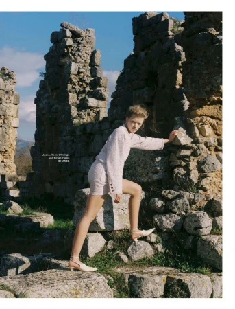 Model posing on ancient stone ruins wearing a textured tweed jacket and shorts with pointed heels against a blue sky