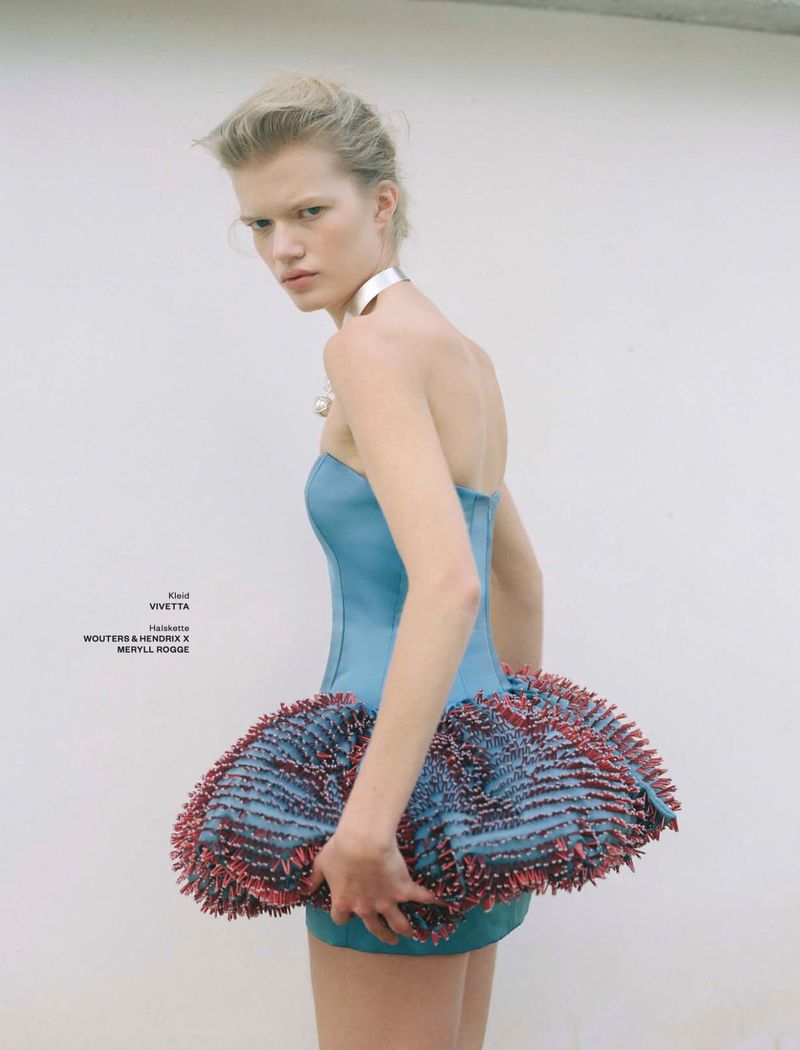 Model wearing a blue structured dress with a ruffled skirt and silver choker against a plain background
