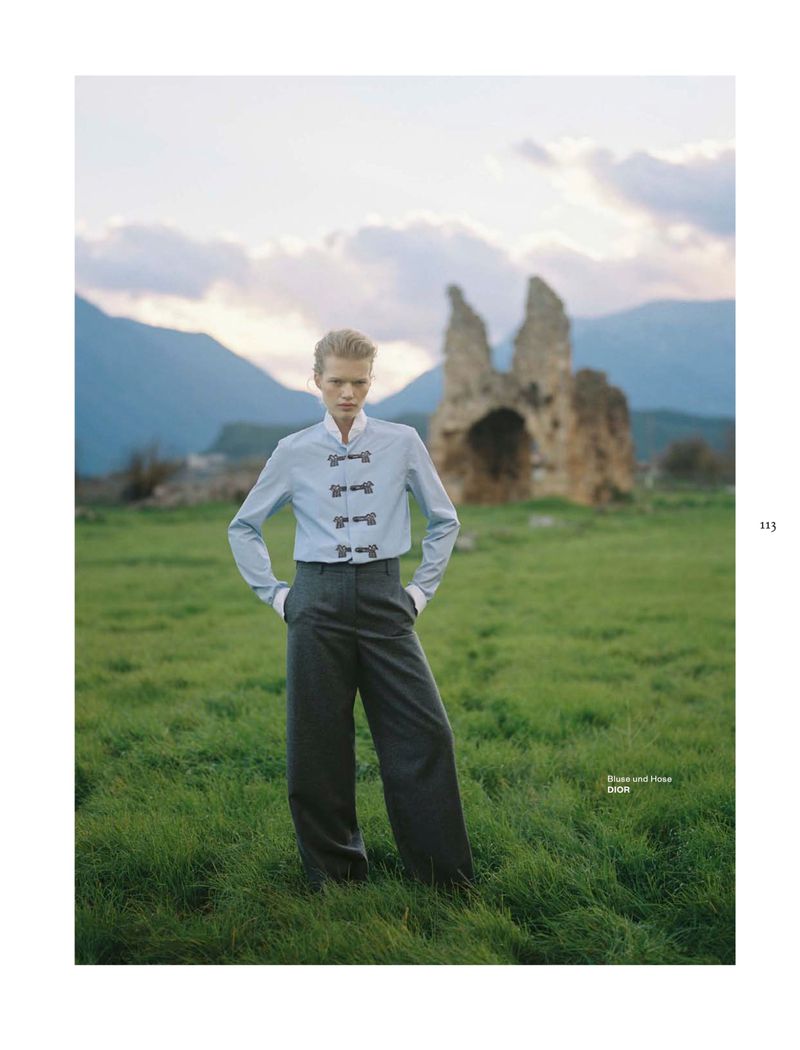 Model standing in a grassy field wearing a light blue button-up shirt and dark grey trousers with mountain and ruin backdrop