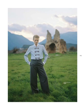 Model standing in a grassy field wearing a light blue button-up shirt and dark grey trousers with mountain and ruin backdrop