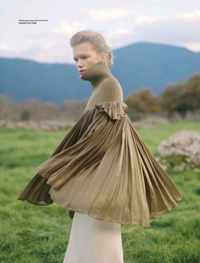 Model wearing a pleated olive green turtleneck sweater in a grassy field with distant rolling mountains under an overcast sky