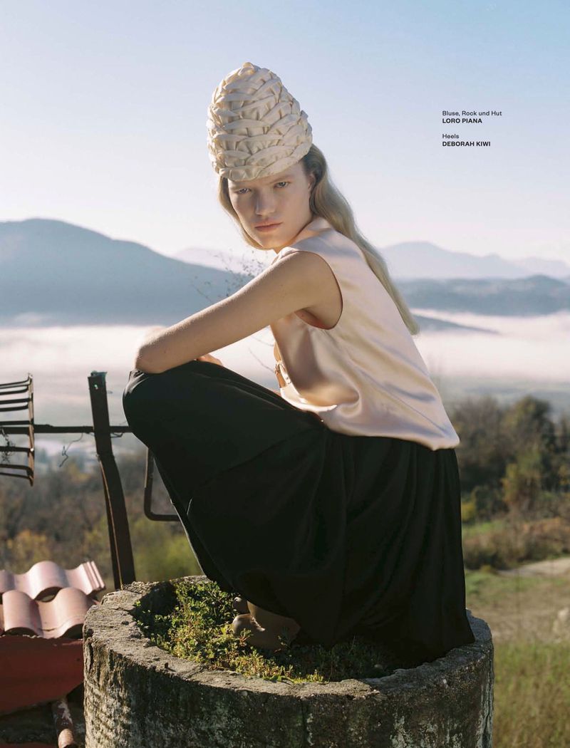 Model crouching outdoors wearing a structured hat, satin top, and dark skirt against rolling hills