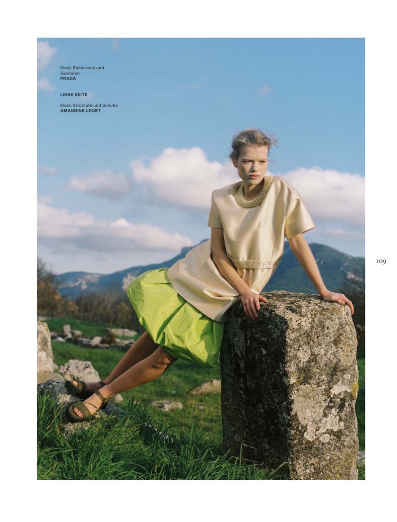 Model seated on a rocky outcrop in a meadow wearing a cream top and lime skirt against mountains under a cloudy sky.