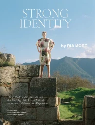 Model standing on ancient stone ruins wearing a structural floral print dress against a mountain landscape under a clear blue sky