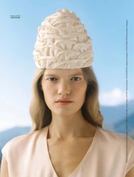 Model wearing a textured cream hat and light-colored top against a blurred mountain backdrop for Flair Germany.