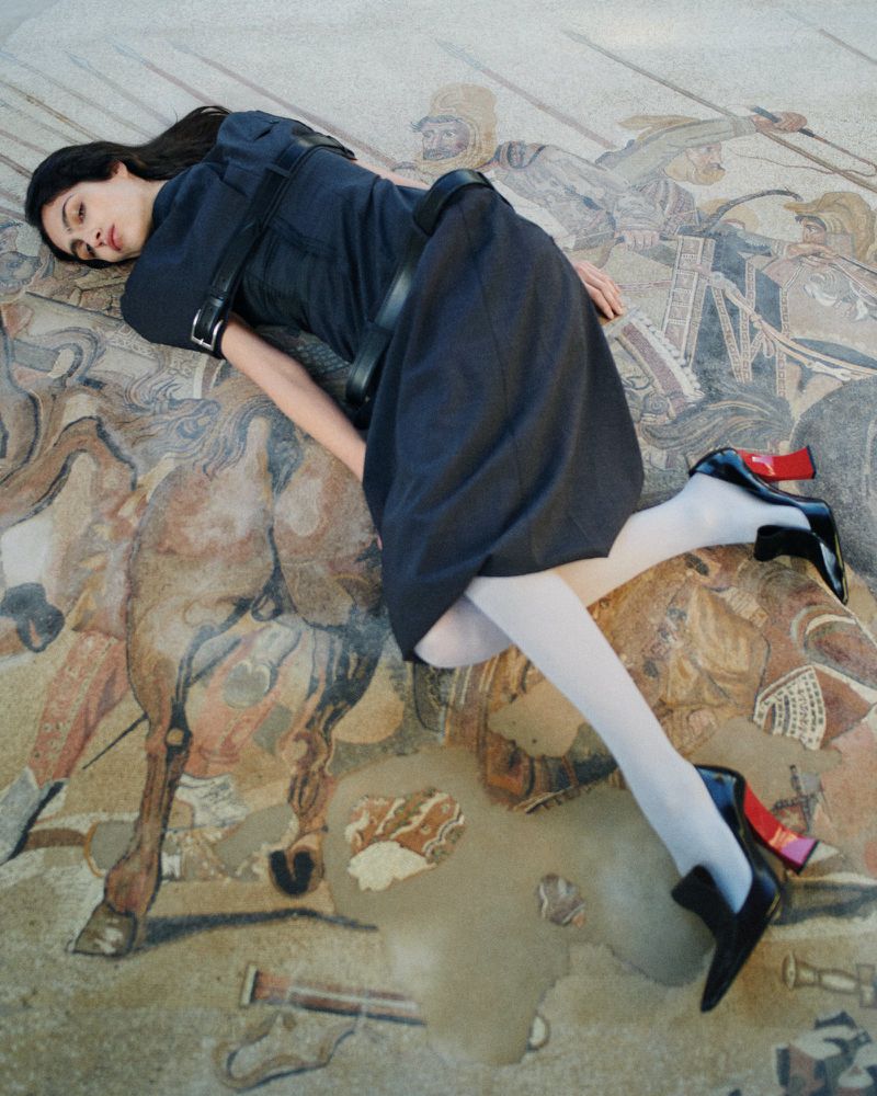 Model lying on an intricate mosaic floor wearing a dark dress, white tights, and black heels