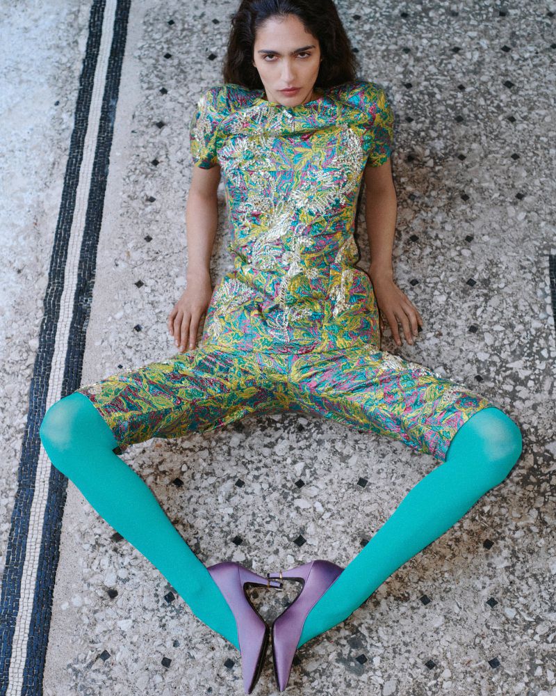 Model seated on a patterned stone floor wearing a green patterned matching set, bright turquoise tights, and purple heels
