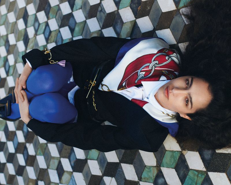 Eye-level view of a stylish woman reclining on a geometric tile floor with long dark hair, wearing a blazer and scarf.