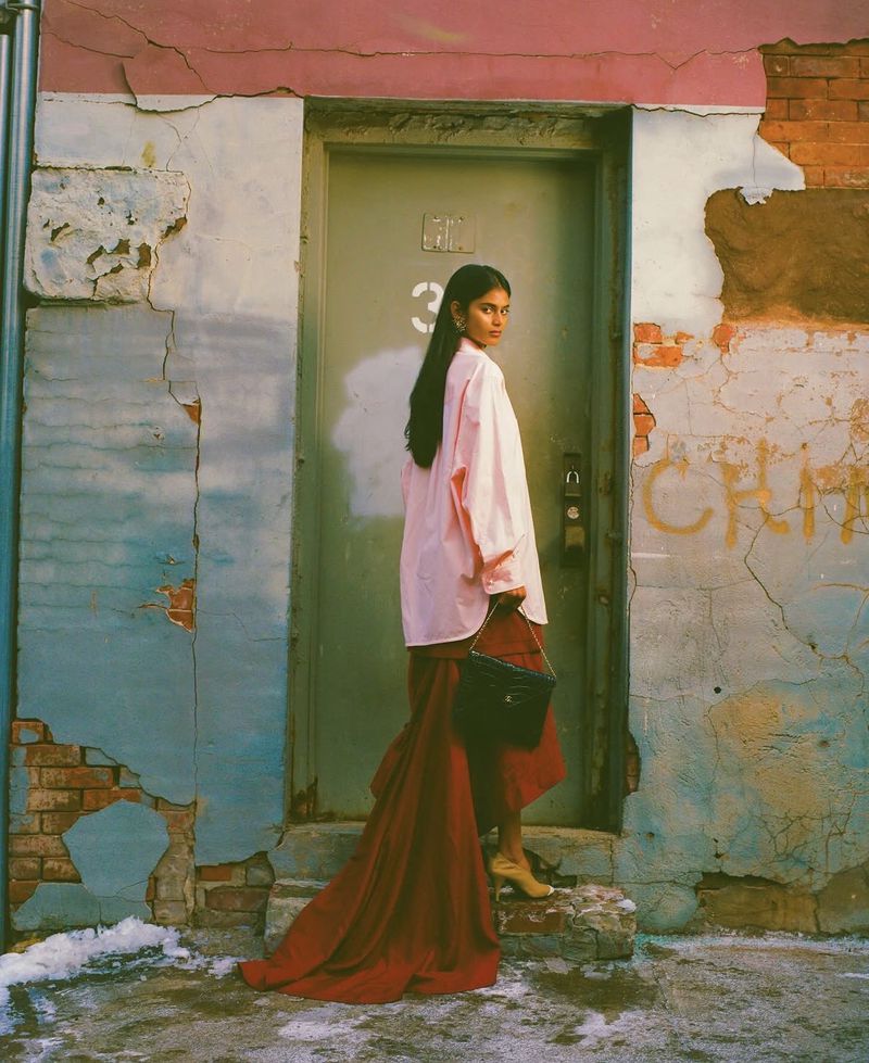 Model wearing a pink shirt and long red skirt holding a handbag standing before a green door on a textured urban street