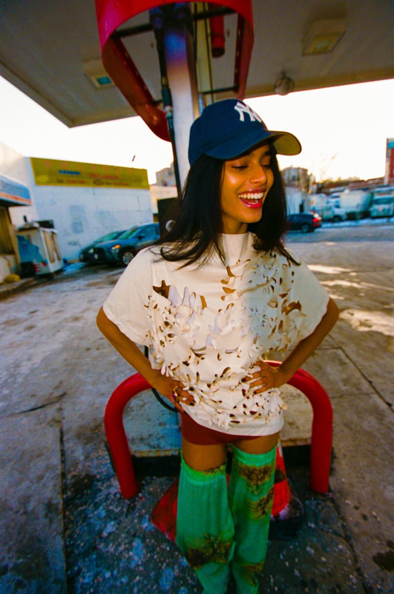 Model Bhavitha Mandava wearing a baseball cap and distressed white top at a service station for The Perfect Magazine.