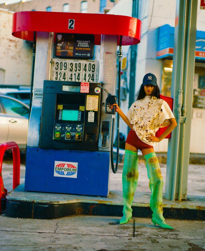 A woman posing at a gas pump wearing a baseball cap, white t-shirt, red shorts, and green boots, standing on a concrete pad.