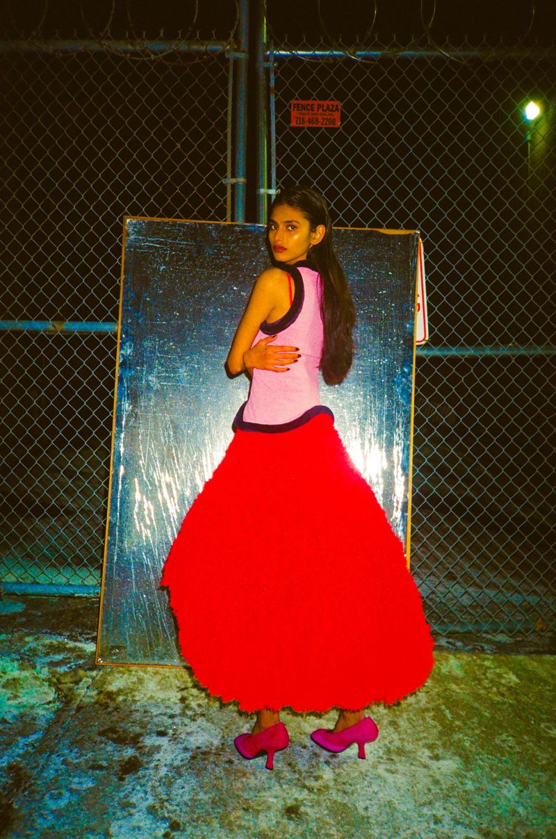 Model Bhavitha Mandava in a pink top and voluminous red skirt posing outdoors at night against a chain link fence for The Perfect Magazine.