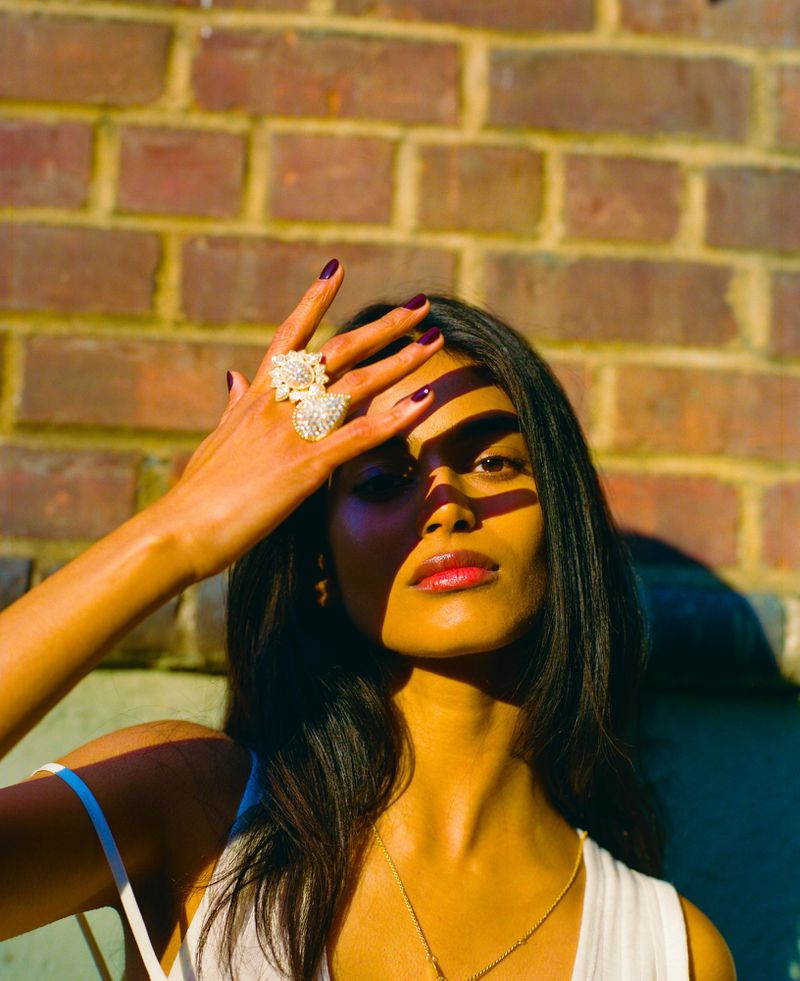 Model Bhavitha Mandava with raised hand wearing large rings against a brick wall for The Perfect Magazine editorial.
