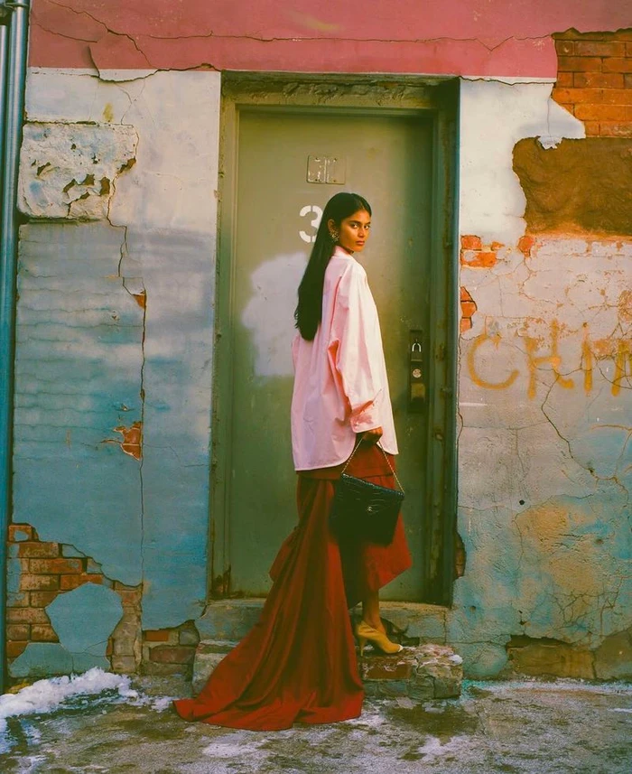 Model wearing a pink shirt and long red skirt holding a handbag standing before a green door on a textured urban street