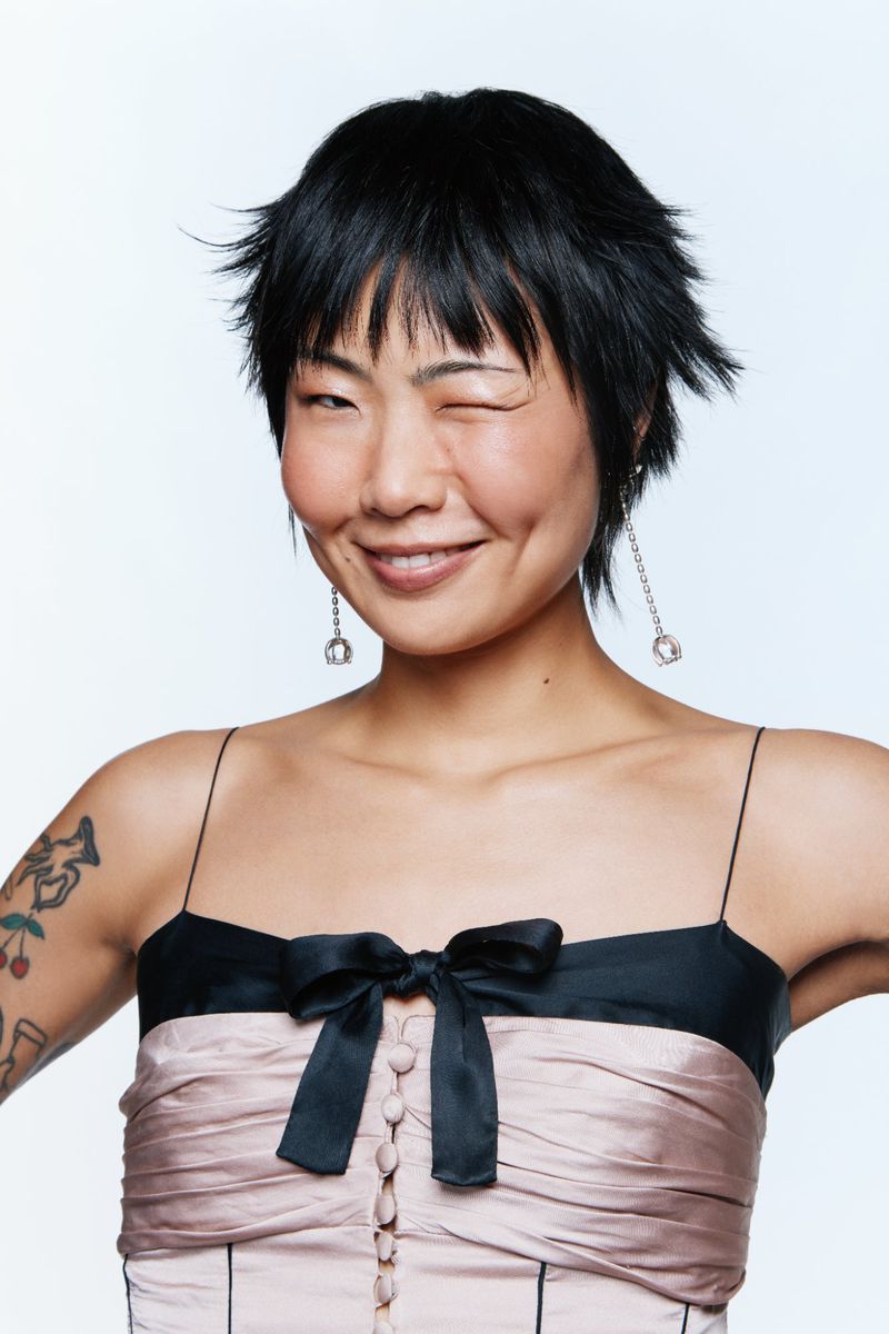 Portrait of model May Hong winking, wearing a strapless pink and black dress with delicate earrings against a white background