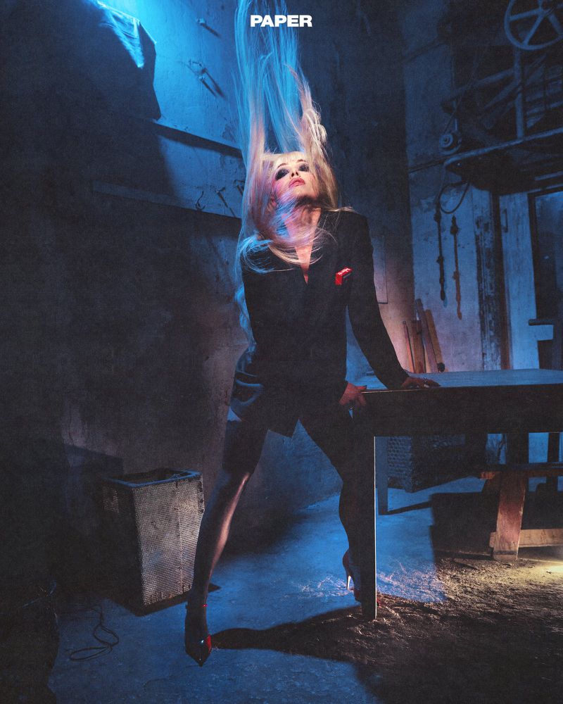 Blonde woman wearing a black jacket posing dramatically with hair flying upward in an industrial room with blue lighting