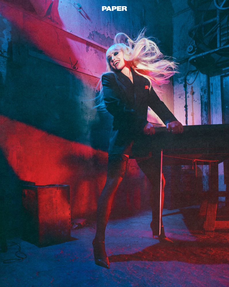 Blonde woman wearing a dark blazer in a gritty industrial space with red and blue lighting for Paper Magazine editorial.
