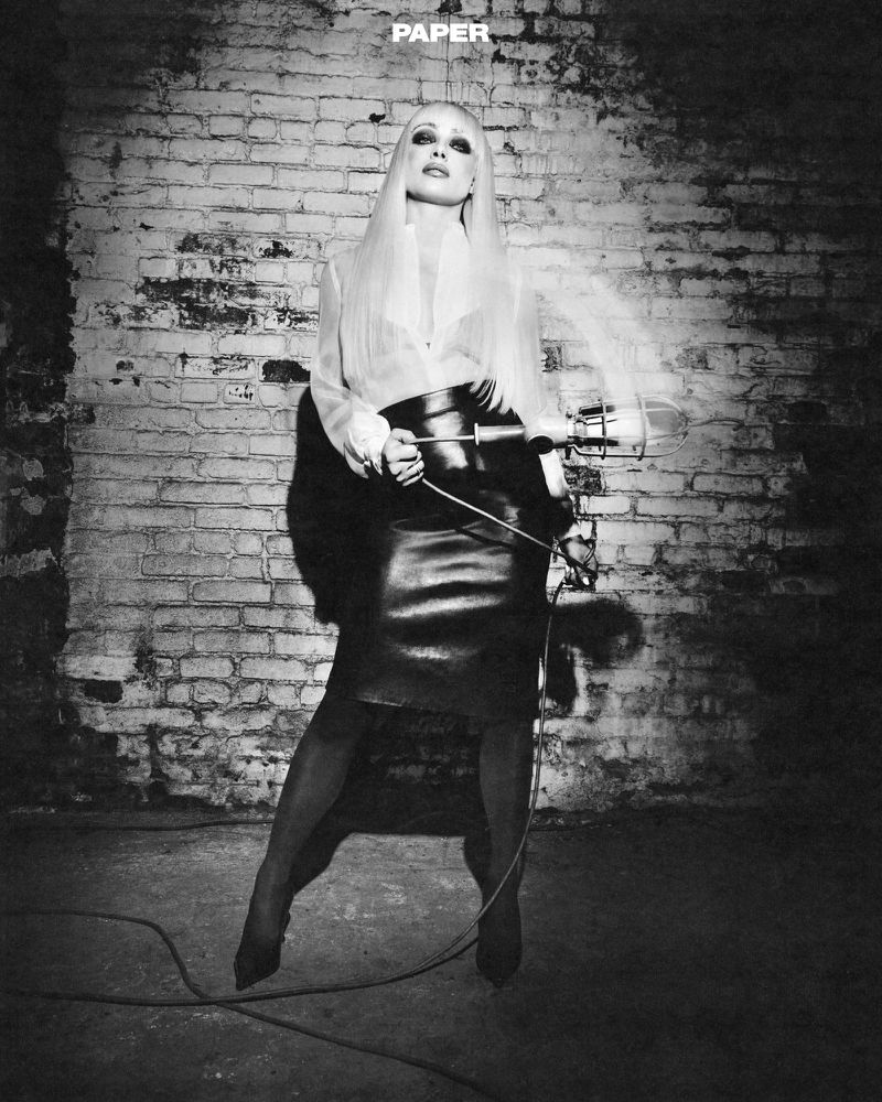 Black and white editorial portrait of a blonde woman holding a work light against an exposed brick wall background.