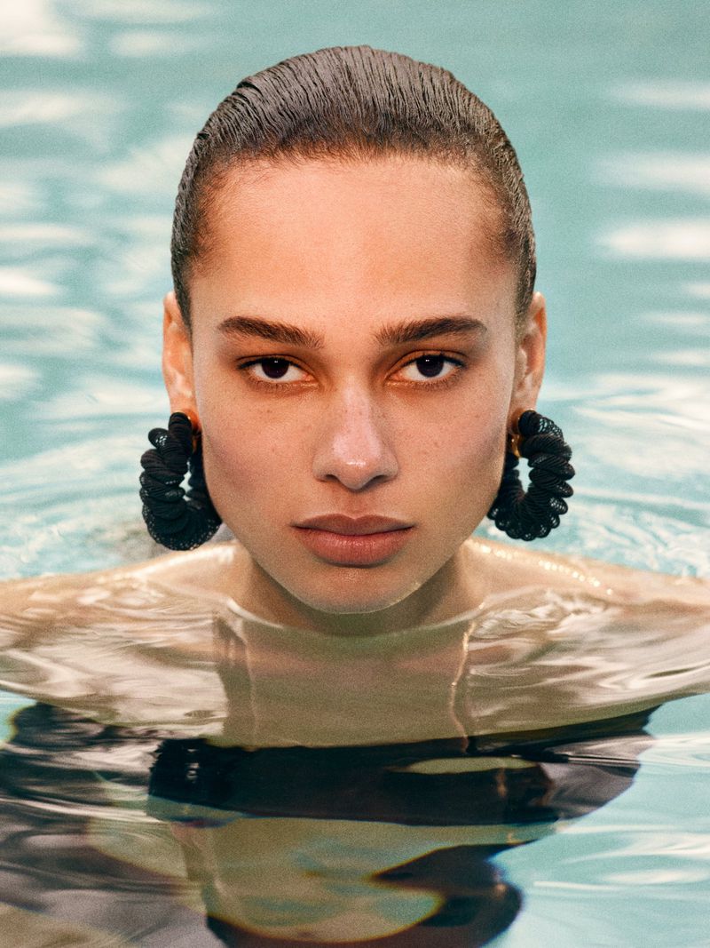 Close-up of a woman with short, dark hair in a pool, wearing black twisted earrings, looking directly at the camera.