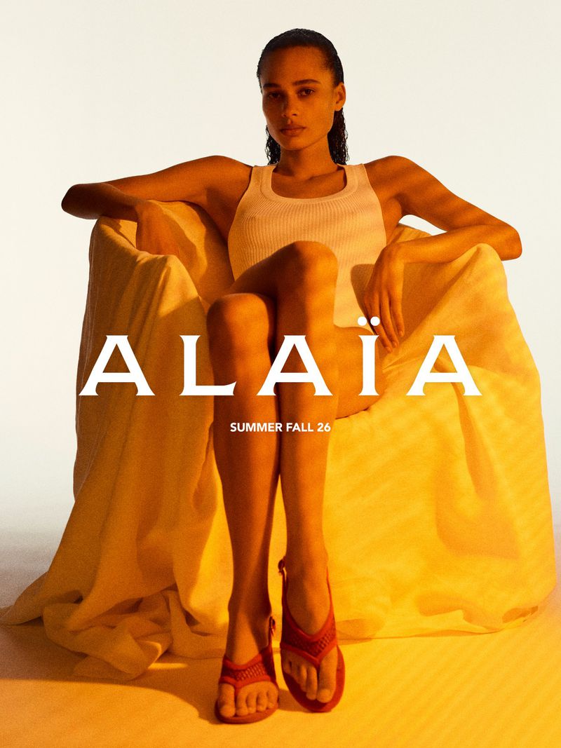 Model seated in an armchair wearing a white tank top and red shoes against a pale background for Alaïa