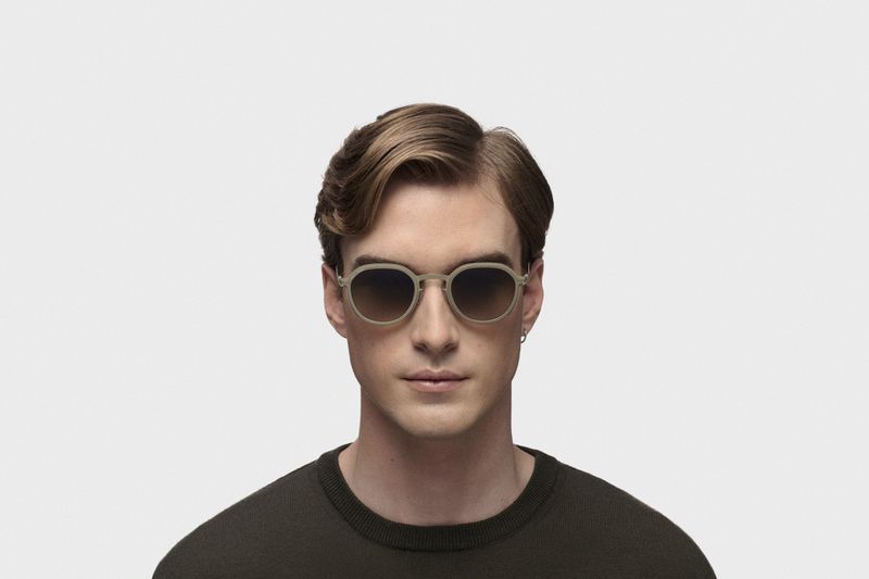 Close-up of a light-skinned adult male wearing round, tan-colored sunglasses and a dark olive green sweater on a white background.