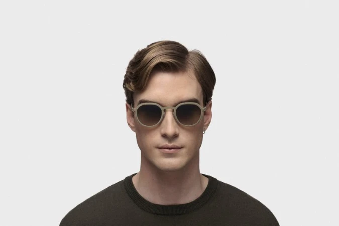 Close-up of a light-skinned adult male wearing round, tan-colored sunglasses and a dark olive green sweater on a white background.