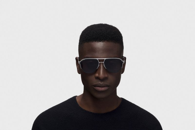 Close-up portrait of a Black man wearing silver aviator sunglasses and a black sweater against a plain white background.