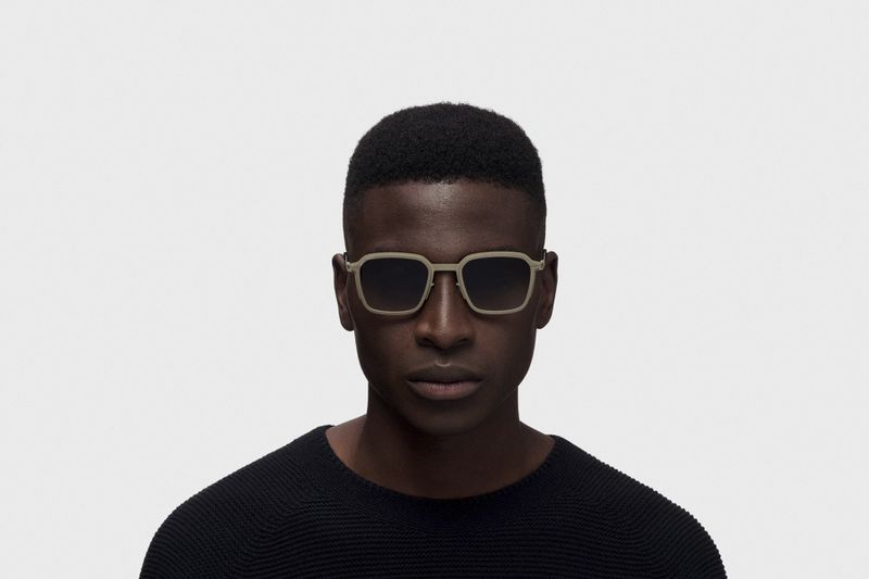 Black man wearing square beige sunglasses and a black sweater against a white seamless background for RIMOWA X MYKITA.