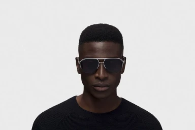 Close-up portrait of a Black man wearing silver aviator sunglasses and a black sweater against a plain white background.