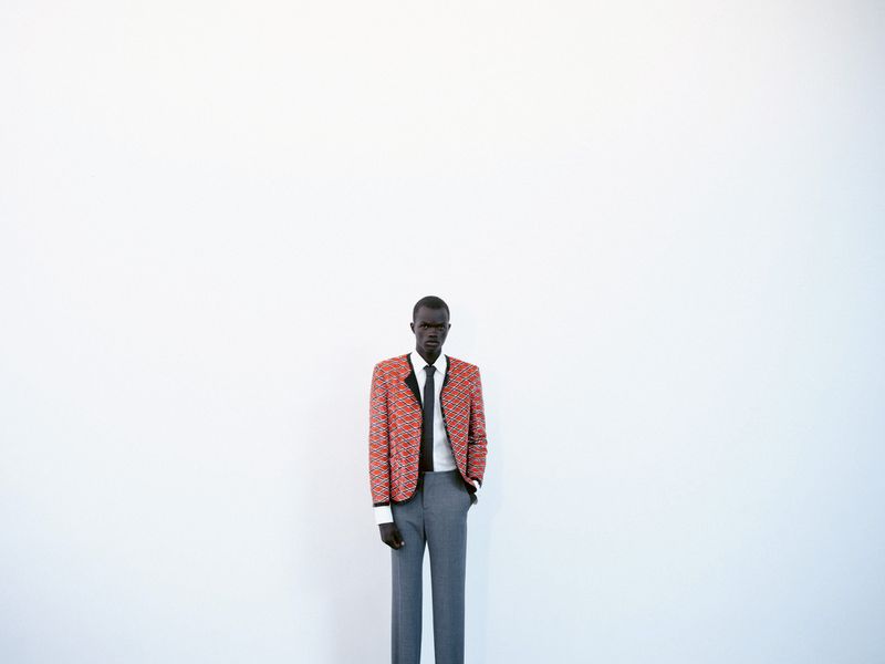 A lone adult Black man stands in an indoor photo studio wearing a patterned blazer, tie, and grey trousers against a white backdrop.