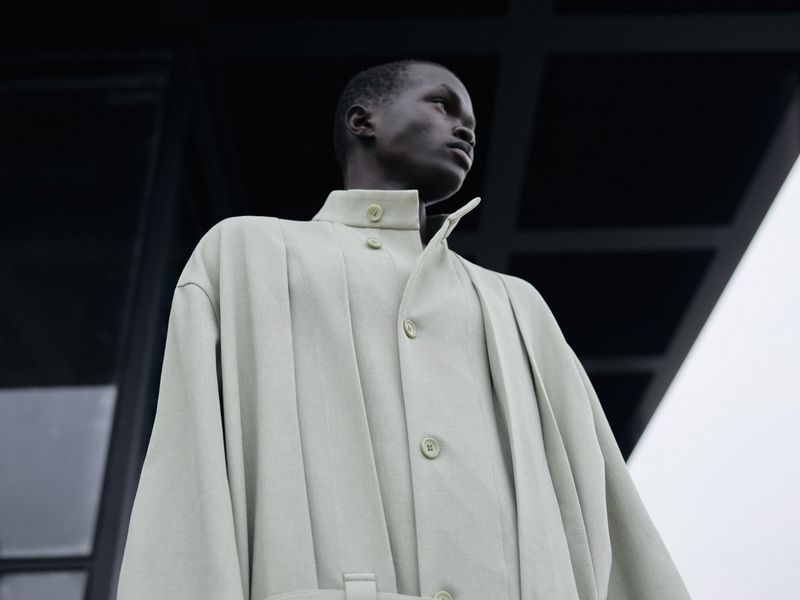 Low-angle shot of a black male model in a light green structured coat against a dark architectural exterior.